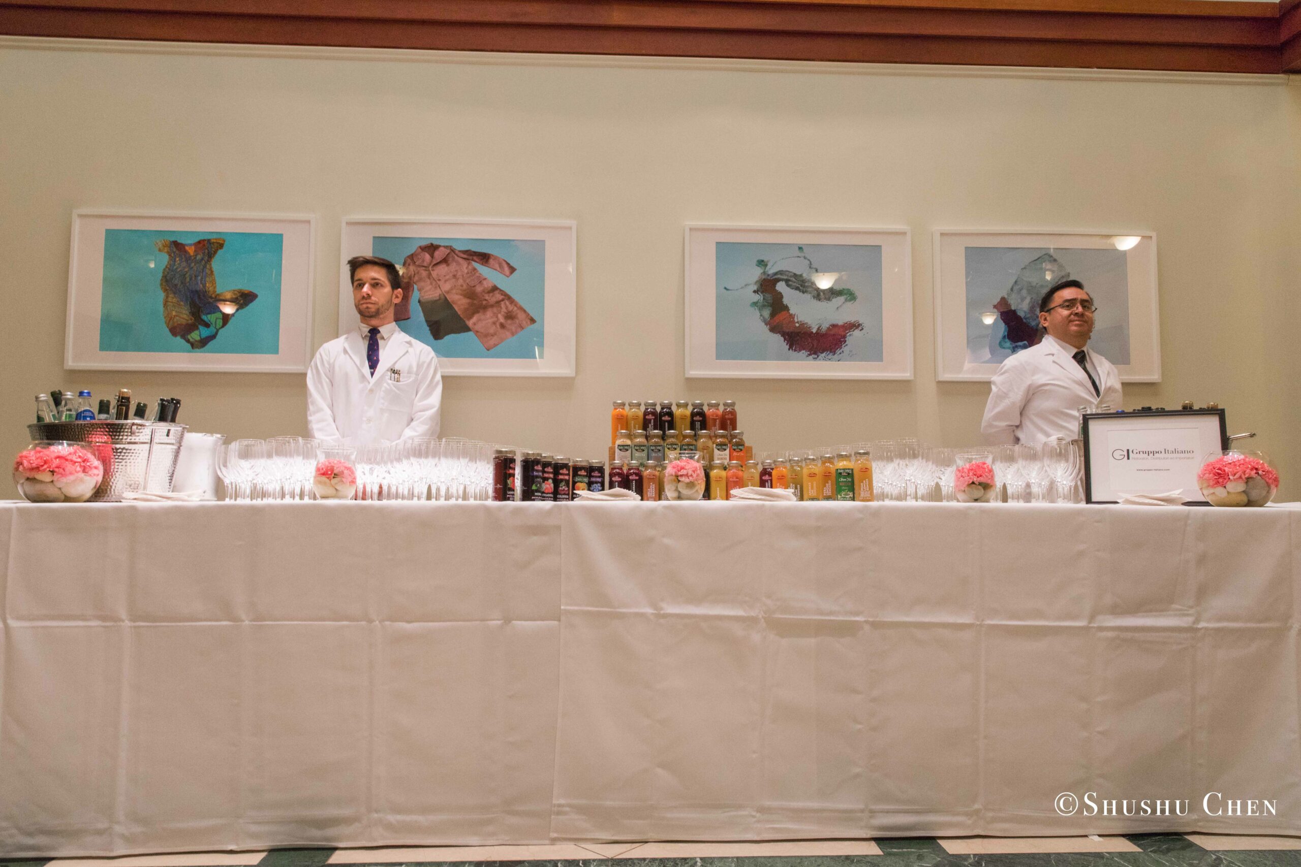 two-bartenders-stand-behind-a-long-table-covered-in-a-white-cloth-fully-equipped-with-glasses-and-bottles-from-Italian-table-talks-at-casa-italiana
