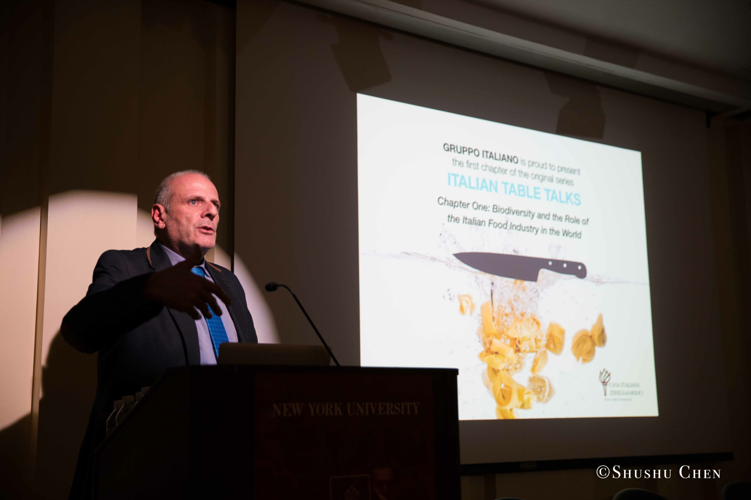 stefano-presenting-at-a-podium-with-a-projector-screen-behind-him-from-Italian-table-talks-at-casa-italiana