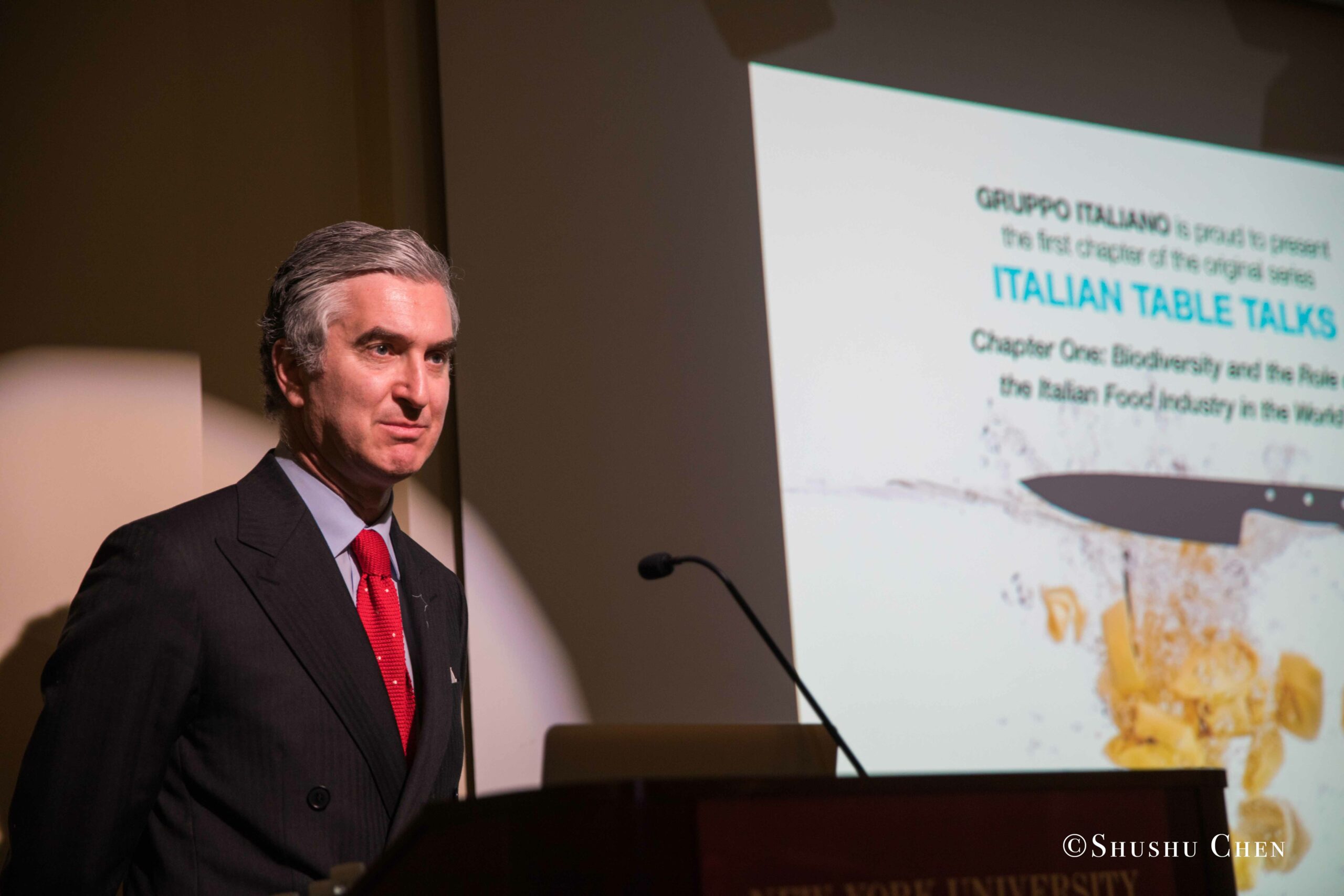 a-man-in-a-suit-stands-at-a-podium-presenting-from-Italian-table-talks-at-casa-italiana