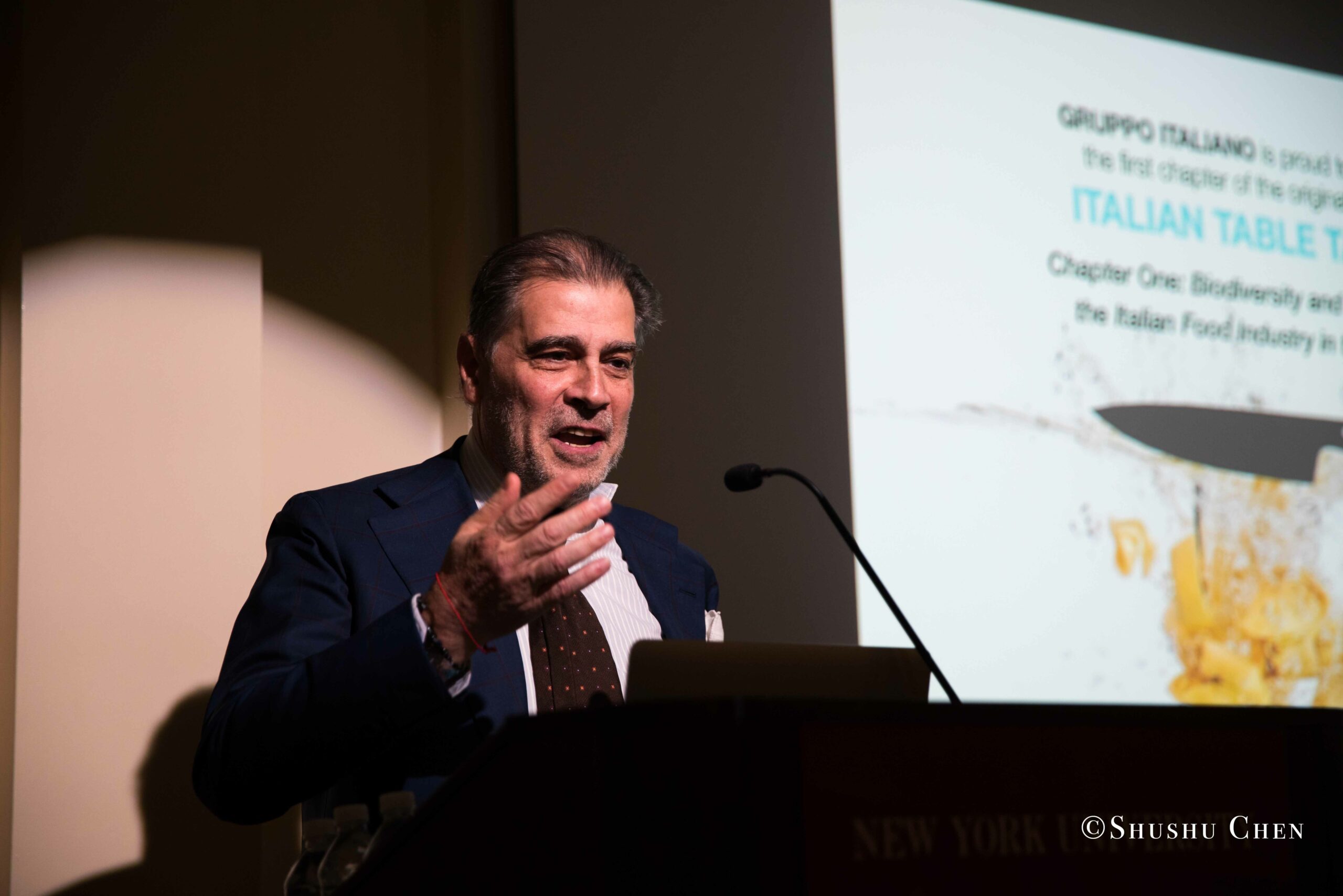 a-man-gestures-while-speaking-at-a-podium-with-a-presentation-screen-in-the-background-from-Italian-table-talks-at-casa-italiana