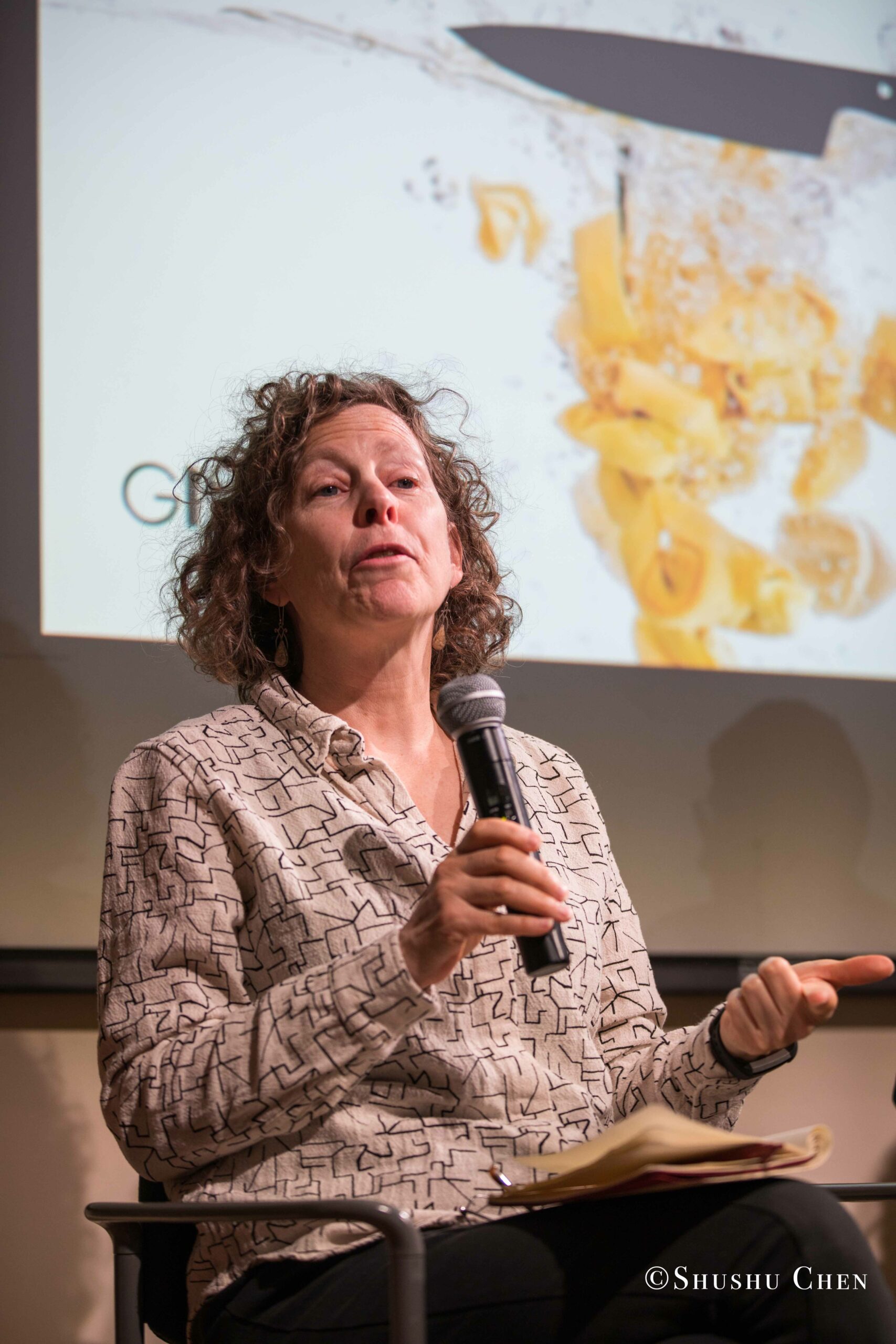 a-woman-with-curly-hair-holding-a-microphone-speaking-from-Italian-table-talks-at-casa-italiana