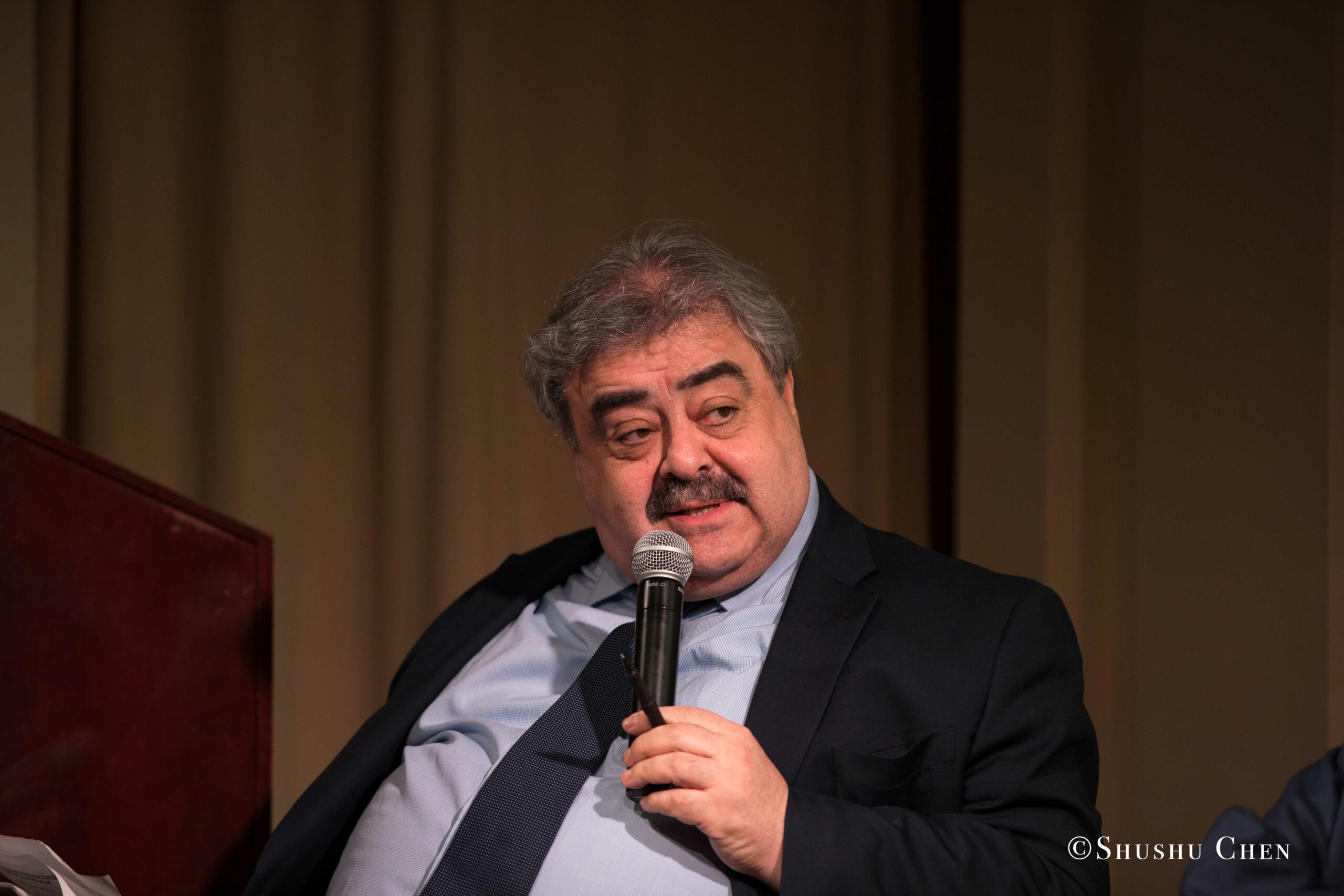 a-man-with-gray-hair-and-a-mustache-wearing-a-dark-suit-and-light-blue-shirt-holds-a-microphone-while-speaking-from-Italian-table-talks-at-casa-italiana