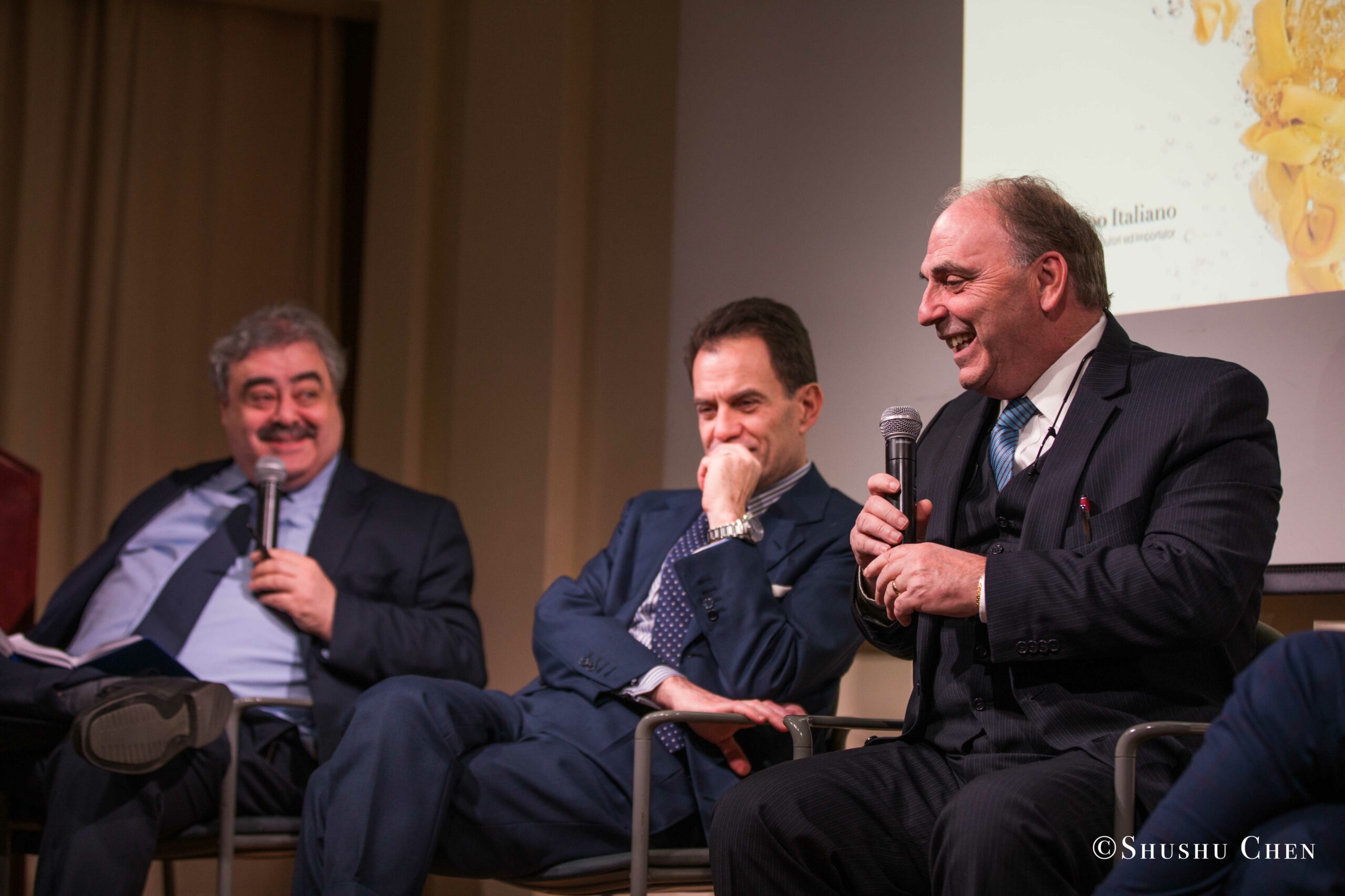 Three-men-in-formal-attire-are-sitting-on-a-stage-with-the-one-on-the-right-speaking-into-a-microphone-and-laughing-while-the-others-listen-one-smiling-and-the-other-deep-in-thought-from-Italian-table-talks-at-casa-italiana