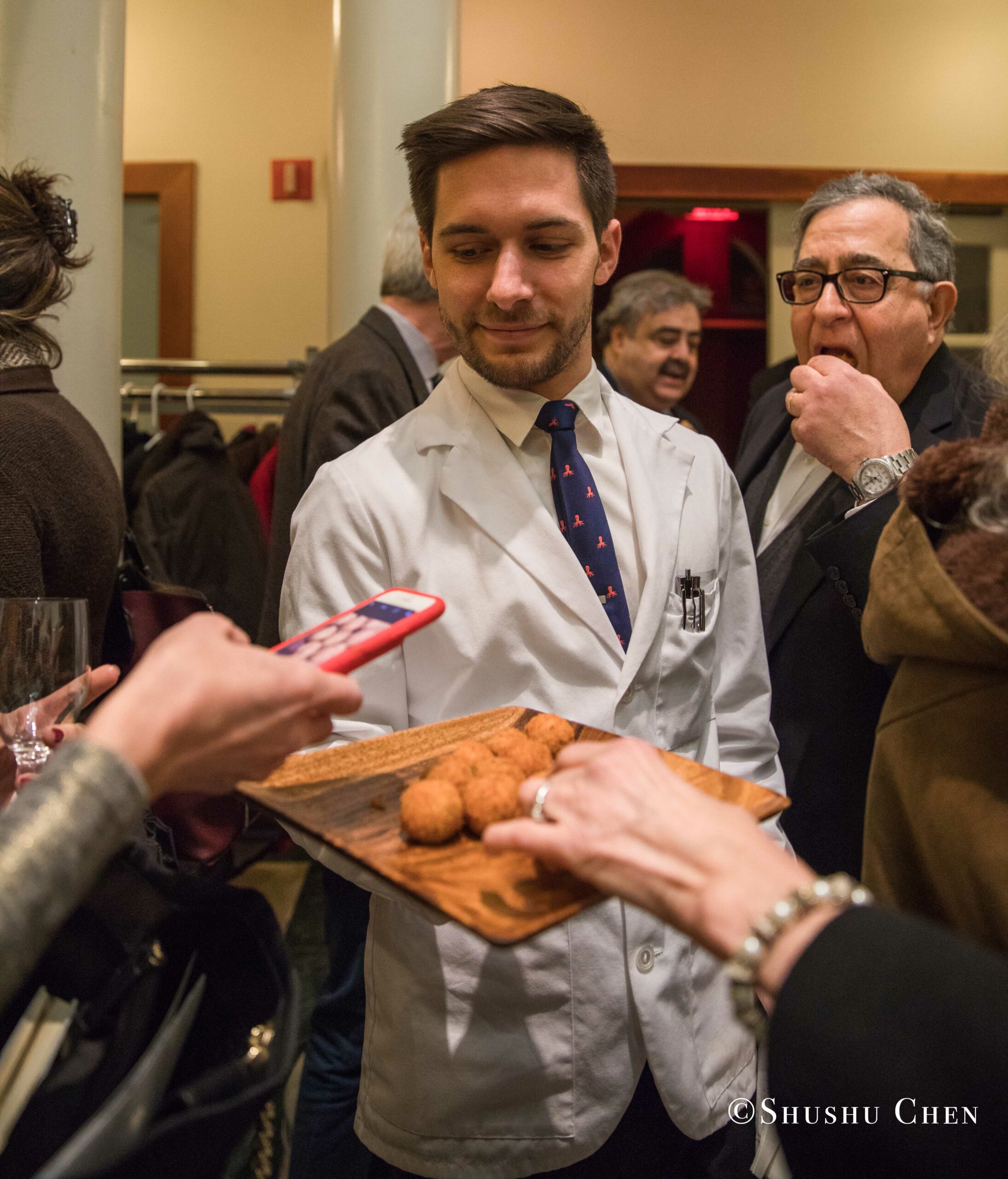 a-man-in-a-white-lab-coat-and-tie-offers-a-tray-of-cookies-to-people-from-Italian-table-talks-at-casa-italiana