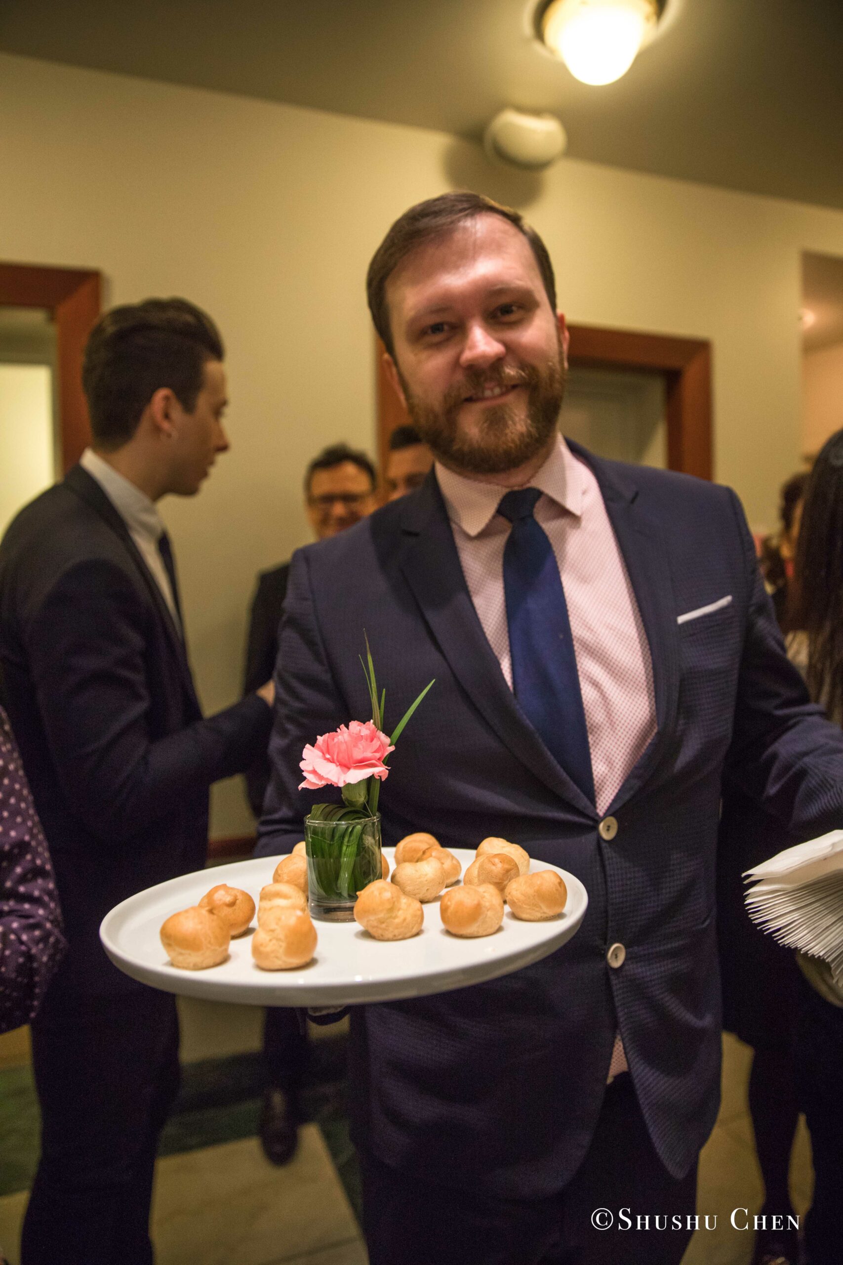 a-male-server-in-a-suit-and-tie-smiling-as-he-offers-a-tray-of-cream-puffs-from-Italian-table-talks-at-casa-italiana