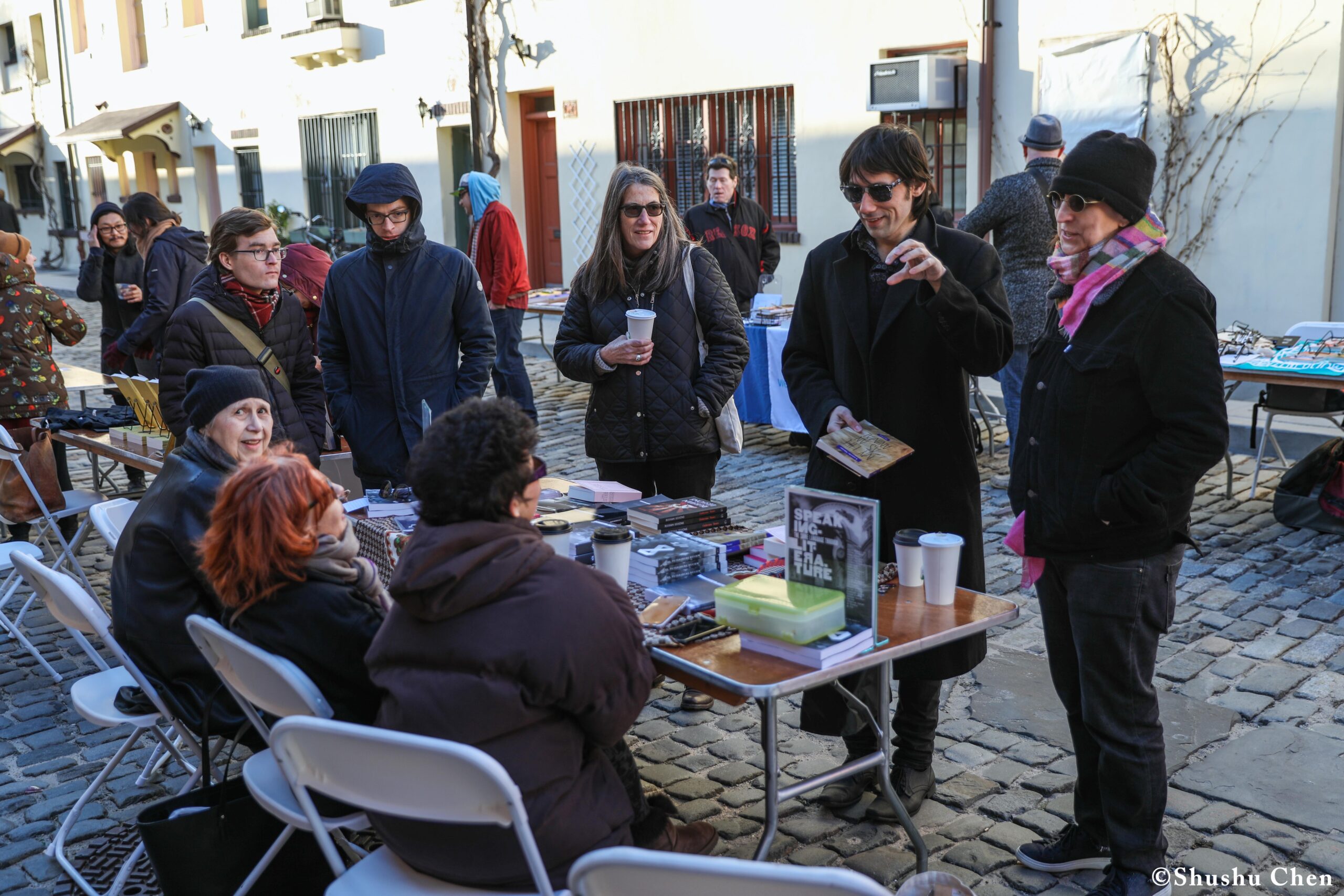 group-of-people-gathered-around-a-table-filled-with-books-at-a-street-market-conversing-and-enjoying-drinks-on-a-sunny-day-from-pen-world-voices-literary-mews-at-nyu