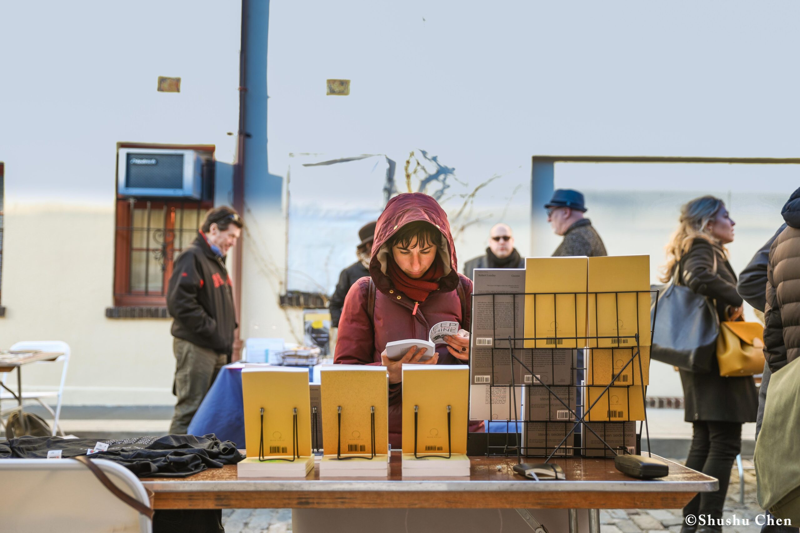 a-woman-in-a-pink-headscarf-intently-reads-a-book-at-an-outdoor-market-stall-from-pen-world-voices-literary-mews-at-nyu