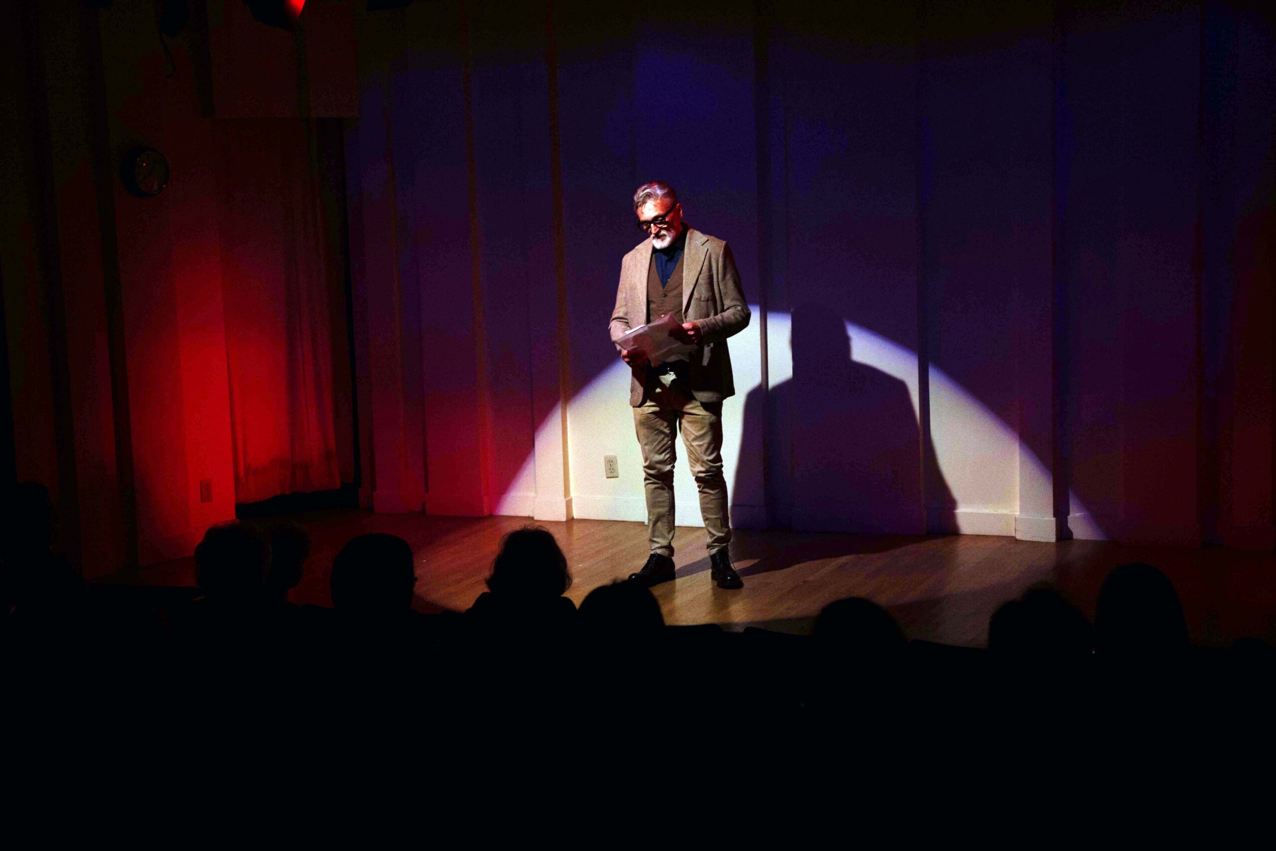 A man stands on stage under a spotlight, holding a piece of paper. He wears glasses, a blazer, and jeans. The background is dimly lit with red and blue lights, and an audience is visible in the shadows.