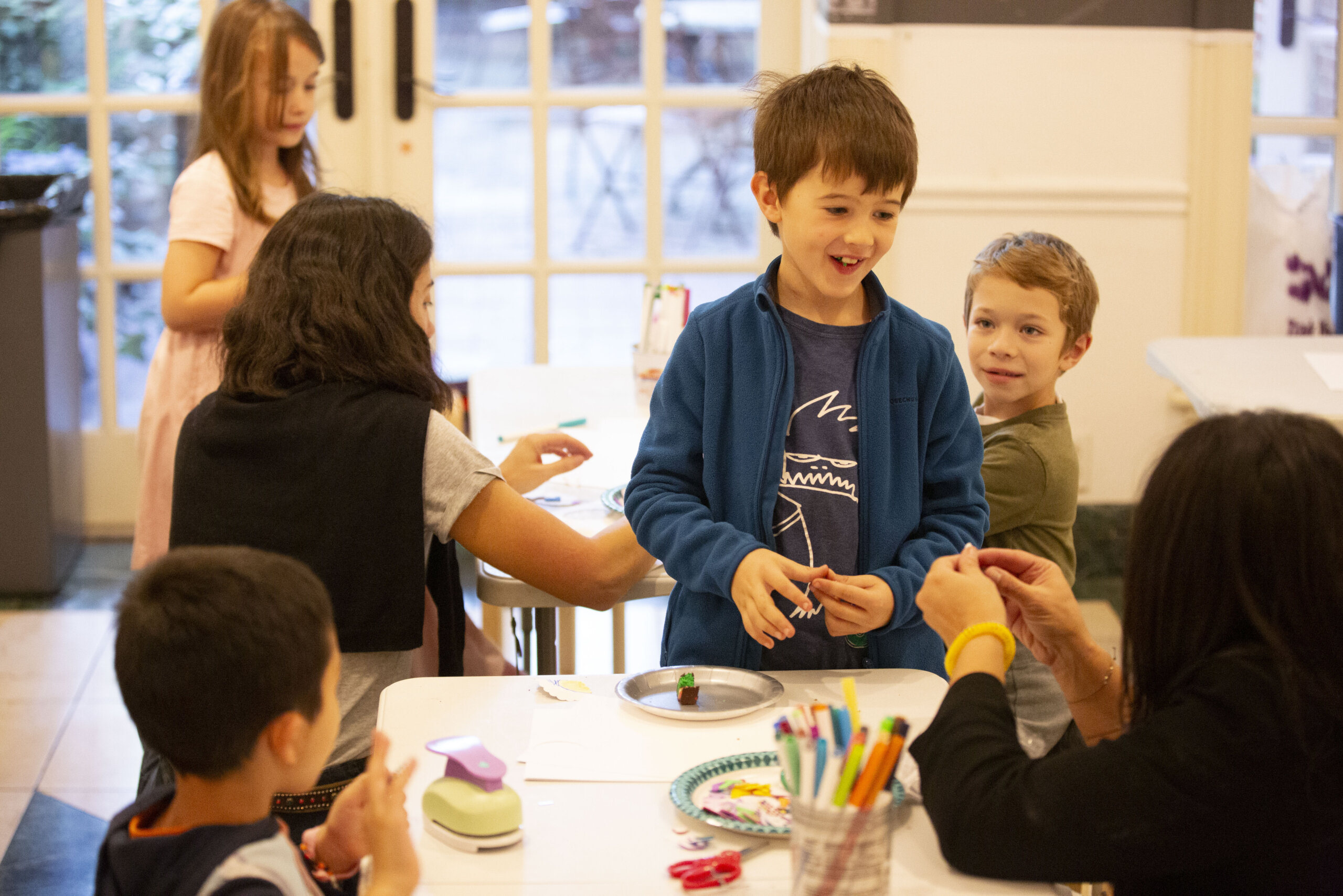 Children are gathered around a table, engaging in arts and crafts activities. One boy is smiling, standing in front of the table, while others focus on their projects. A variety of art supplies are scattered on the table.